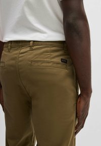 Khaki bukser med en glat tekstur; har to baglommer og et lille sort logo-label nær taljebåndet.