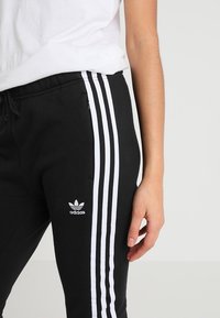 Svarta sweatpants med vitt tre-stripes mönster, med en logotyp på vänster lår, elastisk midja och sidoficka med dragkedja.