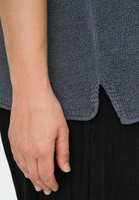 Szary dzianinowy top z fakturowanej tkaniny, z bocznymi rozporkami i luźnym krojem, zestawiony z czarną odzieżą pod spodem. Ręka częściowo widoczna.