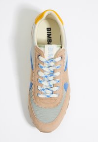 Zapatilla con una base de ante beige, acentos azul claro y paneles de malla. Presenta superposiciones en forma de onda, cordones azules y una tapa de talón amarilla.
