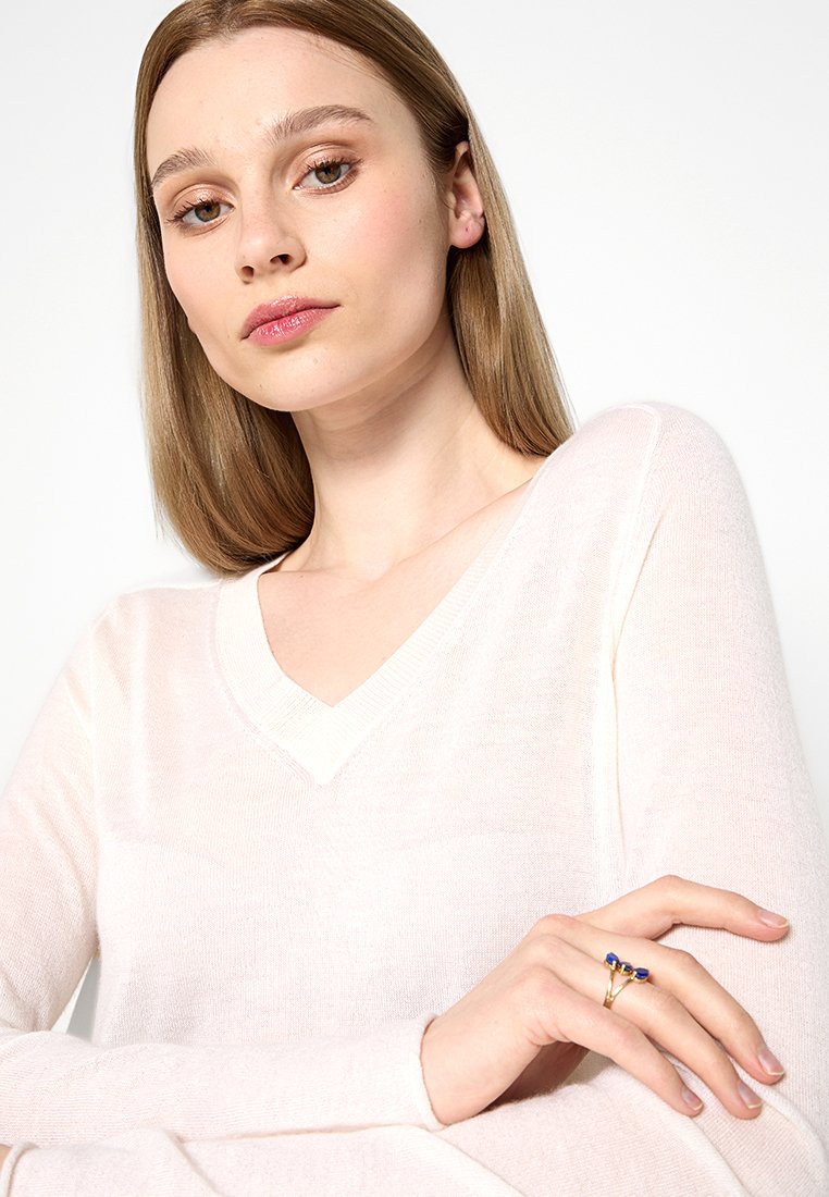 Helles rosa V-Ausschnitt-Pullover aus weichem, leichtem Stoff. Hat lange Ärmel und eine Hand, die mit einem goldenen Ring mit blauen Akzenten geschmückt ist.