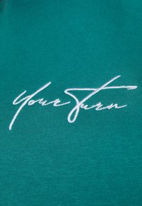 Tela de algodón color azul verdoso con una textura suave que presenta un texto bordado en blanco que dice "Tu turno" en estilo cursivo.