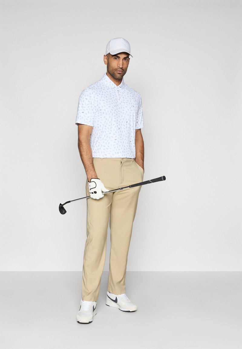 Golfbekleidung mit einem kurzärmeligen blauen gemusterten Polo, beigen Hosen, weißen Sneakers und einer weißen Kappe, während er einen Golfschläger hält.