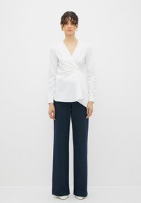Blusa avvolgente bianca con maniche lunghe e arricciatura laterale. Abbinata a pantaloni blu scuro a gamba larga e scarpe bianche a punta. Orecchini a cerchio in oro.