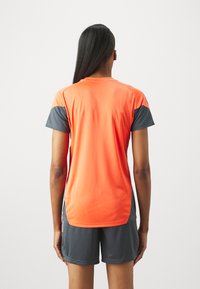 Μπλούζα αθλητικής ένδυσης με κοντό μανίκι σε φωτεινό πορτοκαλί χρώμα με γκρι λεπτομέρειες. Διαθέτει στρογγυλή λαιμόκοψη και άνετη εφαρμογή, κατασκευασμένη από ελαφρύ ύφασμα.