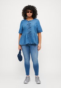 Top peplum de denim azul con mangas cortas, detalle de nudo en el frente, combinado con jeans ajustados azules y zapatillas grises. Sosteniendo un bolso de mano azul oscuro.