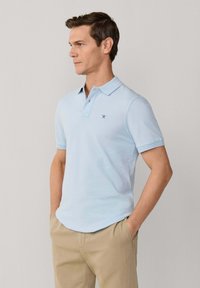 Homme portant un polo à manches courtes bleu clair avec un petit logo, pantalon beige, mains dans les poches, tourné vers la gauche sur un fond uni.