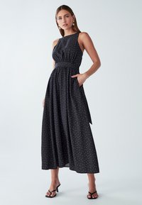 Žena v bezrukávovych čiernych midi šatách s bielymi bodkami, so zvýrazneným pásom, bočnou kapsou a čiernymi sandálmi na podpätku.