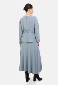 Himmelblaues, langärmliges Kleid mit Peplum-Taille, das einen Rückenschlitz mit Schlüsselöffnung und einen fließenden Rock aufweist, kombiniert mit schwarzen Knöchelstiefeln.