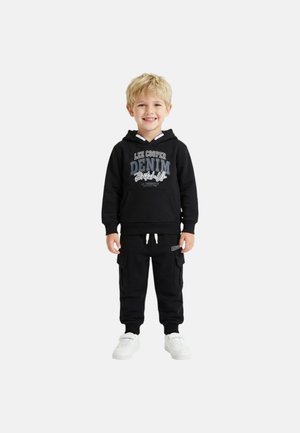 Garçon blond souriant portant un sweat à capuche noir avec le texte "Lee Cooper Denim", un pantalon cargo noir et des baskets blanches, debout sur un fond blanc.