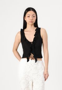 Top negru fără mâneci, scurt, cu decolteu adânc în V și margini volanate, având o închidere cu fundiță în față. Purtați-l cu pantaloni albi cu model din dantelă.