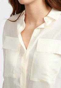 Blusa leggera e trasparente di colore crema con colletto, due tasche frontali e chiusura a bottoni. Il tessuto presenta una superficie liscia e leggermente traslucida.