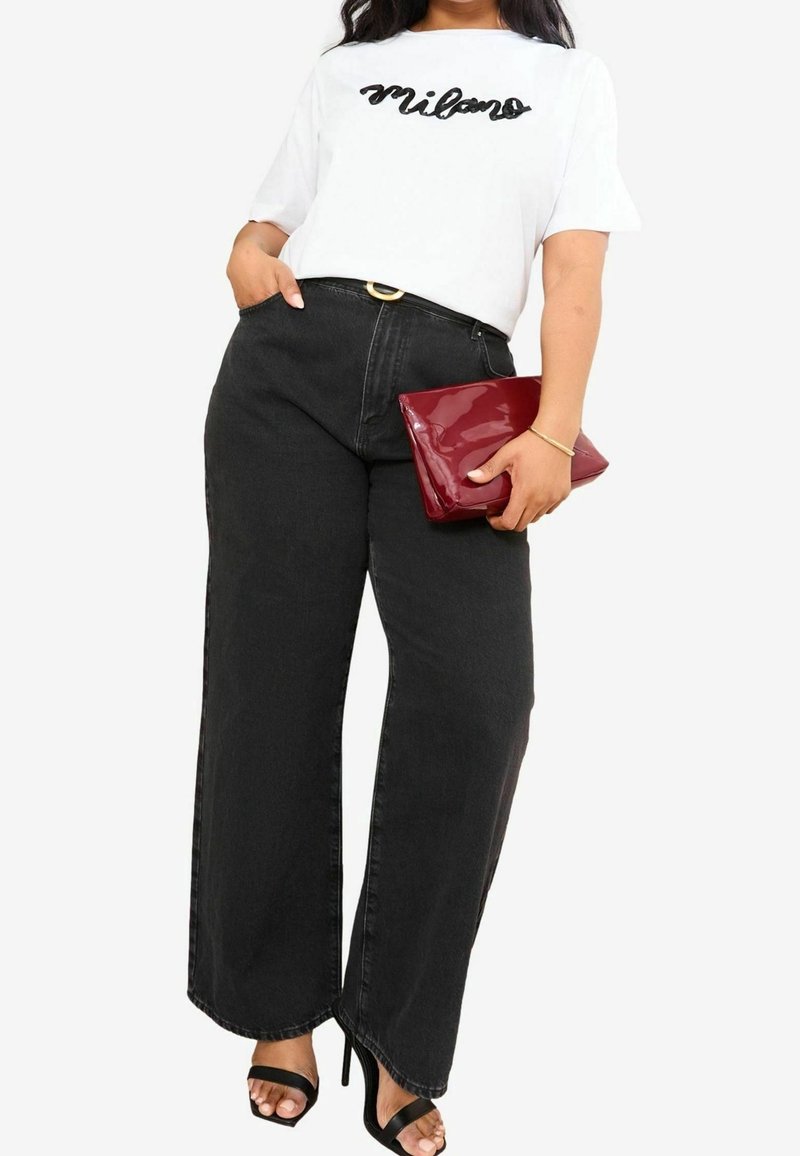 Nainen pukeutuneena valkoiseen "Milano"-t-paitaan, korkeavyötäröisiin mustiin leveälahkeisiin farkkuihin, mustiin korollisiin sandaaleihin ja kädessään kiiltävä marjapuuronpunainen kirjekuorilaukku.