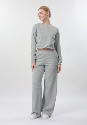 Donna in piedi con maglia grigia a maniche lunghe e pantaloni a gamba larga, indossa sneaker bianche, con i capelli raccolti e lo sguardo di lato.