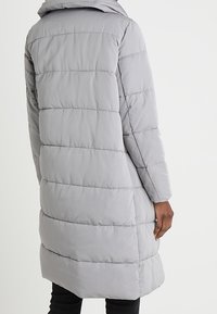Šedý prošívaný kabát s kapucí, s vertikálním prošitím a střední délkou. Viditelná je hladká textura a boční kapsy.