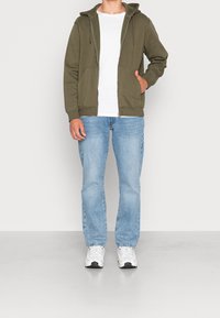 Olivgrüner Reißverschluss-Hoodie mit zwei Fronttaschen, getragen über einem weißen T-Shirt und kombiniert mit hellblauen Jeans und weißen Sneakers.