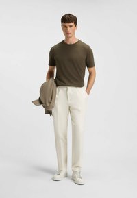 Herreoutfit med en tætsiddende kortærmet skjorte i olivengrøn, skræddersyede hvide bukser og lyse sneakers, med en beige jakke over armen.