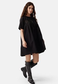 Black cotton dress with short sleeves, round neckline, gathered bodice featuring embroidered accents; paired with knee-high black leather boots.