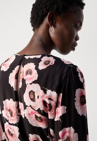 Vestido negro con estampado de grandes amapolas rosas, textura suave, escote redondeado y detalle de ojal en la espalda. Se llevan pendientes de aro plateados.