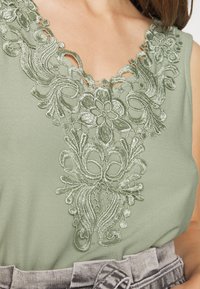 Blusa verde-claro com padrões florais bordados intrincados em um tom ligeiramente contrastante, com decote em V e tecido texturizado.