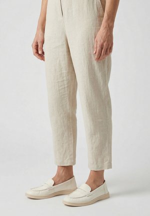 Beige Leinenhose getragen mit off-white Slippern an einer Person, die vor einem neutralen Hintergrund steht.