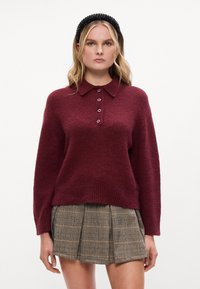 Burgundská pletená mikina s golierom a piatimi gombíkmi, v kombinácii s plisovanou sukňou v kockovanom vzore v neutrálnych odtieňoch a čiernym čelenkou.