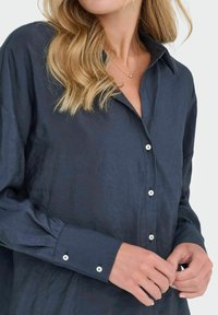 Donna che indossa una camicia button-up blu scuro, slacciando un bottone, con capelli biondi ondeggianti fino alle spalle e una collana d'oro.