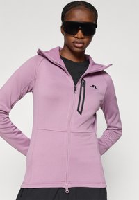 Chaqueta ajustada con cremallera en púrpura claro, hecha de una tela elástica. Presenta un bolsillo con cremallera negro y un acento de logo en el pecho. Diseño con capucha.