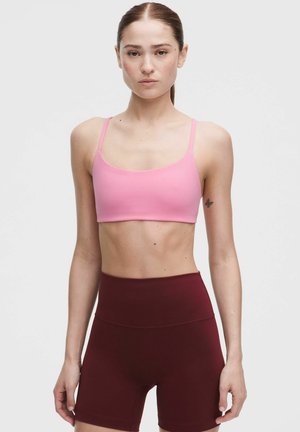 Junge Frau trägt einen pinken Sport-BH und hoch taillierte burgunderfarbene Trainingsshorts, steht mit entspannten Armen vor einem einfarbigen Hintergrund.