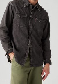 Uomo che indossa una camicia di jeans nera con bottoni e tasche sul petto, pantaloni verde oliva, mano in tasca e tatuaggio visibile sul polso.