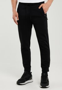 Mees, kes kannab musti dressipükse lukuga taskuga ja musti spordijalatseid, seisab ühevärvilise tausta ees.