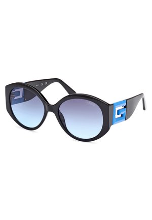 Occhiali da sole neri con una forma a occhio di gatta drammatica, lenti sfumate e un audace logo G blu sul lato. Montatura in plastica liscia.