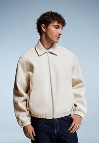 Jachetă din fleece bej cu fermoar în față, guler, manșete și tiv ribbate. Modelul poartă blugi negri, pe un fundal albastru deschis.