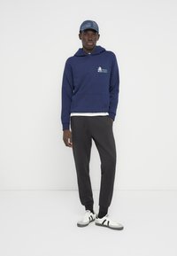 Μπλε ναυτικό hoodie με μπροστινή τσέπη και γραφικό σχέδιο, σε συνδυασμό με γκρι φόρμα και λευκά αθλητικά παπούτσια με μαύρες ρίγες. Τζιν καπέλο baseball.