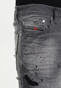 Šedé džíny z denimu s distressed povrchovou úpravou, s několika roztrženinami, stříbrnými doplňky a malou červenou značkovou nášivkou na zadní kapse.