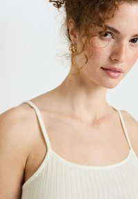 Débardeur côtelé beige clair avec des bretelles fines, présentant un col arrondi et un tissu texturé. Des boucles d'oreilles en or ajoutent une touche d'accent.