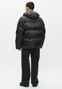 Chaqueta negra llena de plumas con capucha, que presenta secciones acolchadas y una textura lisa y brillante. Combinada con pantalones de pana negros y calzado oscuro.