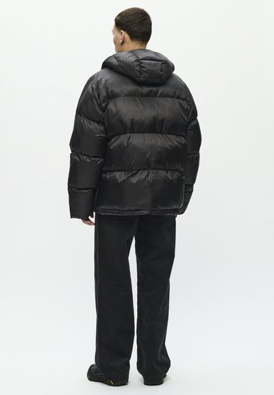 Veste noire rembourrée avec capuche, présentant des sections matelassées et une texture lisse et brillante. Associée à un pantalon en velours côtelé noir et des chaussures foncées.