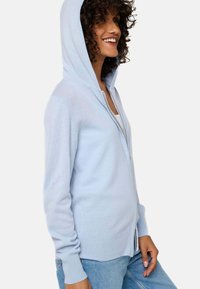 Haut de sweat à capuche zippé bleu clair en tissu doux ; comprend une capuche avec cordon de serrage, des poignets côtelés et des poches latérales, avec une silhouette ajustée.