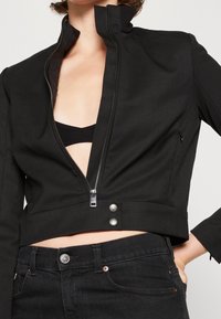 Svart kort jacka med hög krage, framzipper och två silverfärgade tryckknappar i midjan. Slätt tyg med en åtsittande design.