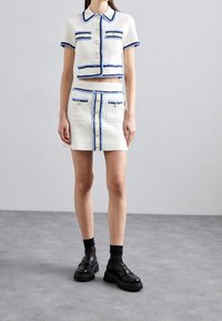 Witte textuur tweedelige outfit bestaande uit een cropped button-up top en een bijpassende mini rok met blauwe rand en accenten op de zakken en knopen.