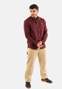 Camicia a maniche lunghe color marrone scuro con righe bianche verticali, abbinata a pantaloni cargo beige e sneakers nere con dettagli arancioni.