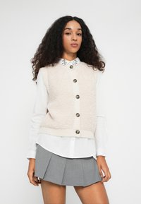 Kvinne med langt krøllete hår iført en hvit skjorte med dekorert krage, beige vest med knapper og grå plissert skjørt som står foran en ensfarget bakgrunn.