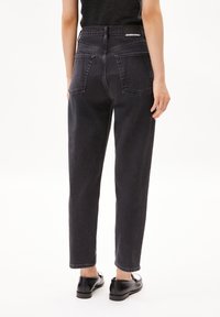 High-waisted black denim jeans with a straight leg design, featuring two back pockets and a logo tag at the waistband. Flat shoes worn.
