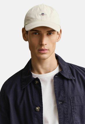 Junger Mann mit beiger Baseballmütze mit Logo, weißem T-Shirt und dunkelblauem, geknöpftem Hemdjacke, der auf einem hellen Hintergrund nach vorne schaut.