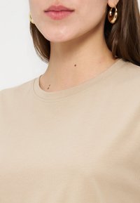 Γυναίκα που φοράει μπεζ μπλούζα με στρογγυλή λαιμόκοψη και μεγάλα χρυσά κρίκους σκουλαρίκια, που δείχνει τον λαιμό και το κάτω μέρος του προσώπου της.