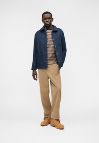 Μπουφάν από μπλε μαρινέ μάλλινο ύφασμα με κλείσιμο με κουμπιά και δύο μπροστινές τσέπες, σε συνδυασμό με καφέ πουλόβερ με σχέδιο και μπεζ παντελόνι κοτλέ. Καφέ μπότες.