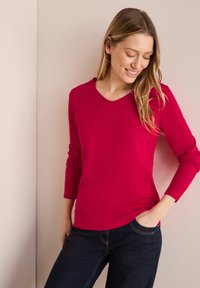 Haut à manches longues et col en V en tissu rose vif. Présente un design ajusté avec des poignets côtelés. Associé à un jean bleu foncé.