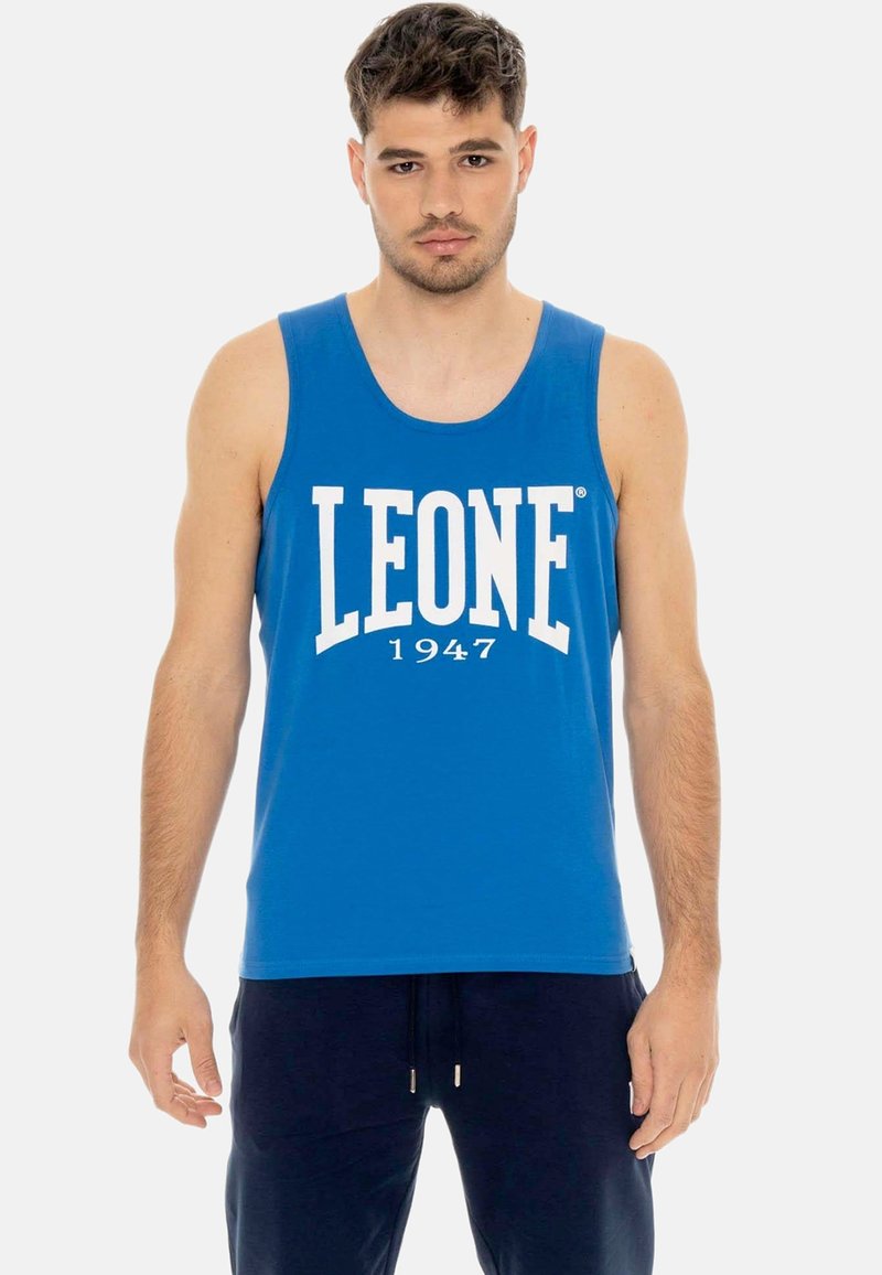 Jeune homme portant un débardeur bleu avec l'inscription « LEONE 1947 » en blanc, debout devant un fond clair uni.