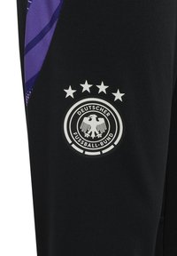 Czarne spodnie sportowe wyposażone w duże białe okrągłe logo z orłem i pięcioma gwiazdami, z fioletowymi akcentami po bokach.